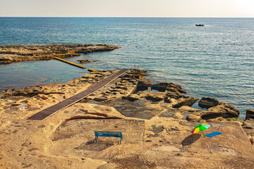 Sunny rocky shore with wooden pier, blue bench, and colorful umbrella by the sea. Summer evening on a deserted natural beach with ladders and rock pools. Travel and relaxation concept.