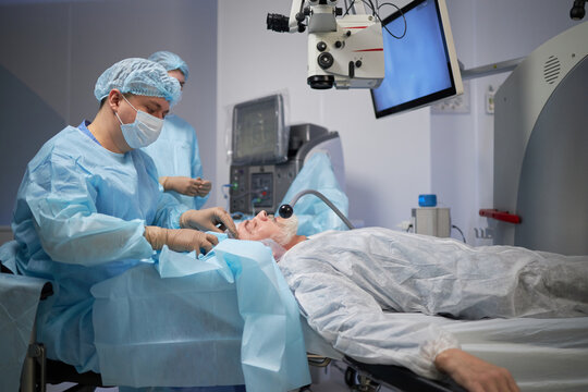 senior man lying on operating table undergoing eye surgery performed by male surgeon wearing surgical mask and gown in modern operating room - Powered by Adobe