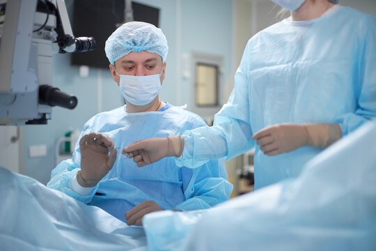 middle aged male surgeon preparing for operation with assistance from young adult female nurse in operating room, both wearing surgical masks and gowns, handling medical instruments