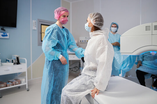 woman sitting on operating table wearing surgical gown and cap, female medical professional standing beside her preparing for procedure in operating room