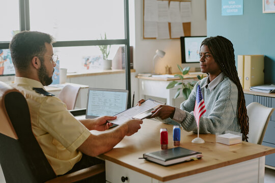 Officer handing documents to woman seated across from desk in office with American flag on display. Natural light coming in through window, illuminating workspace