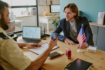 Man in uniform discussing immigration process with young Caucasian man in office setting with...