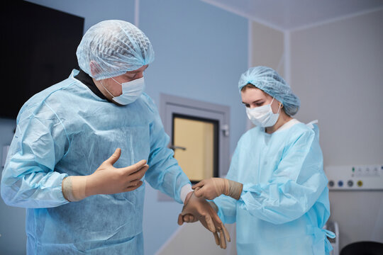 Middle aged man preparing for surgery with woman assisting by putting on sterile gloves in operating room, both wearing surgical masks and gowns