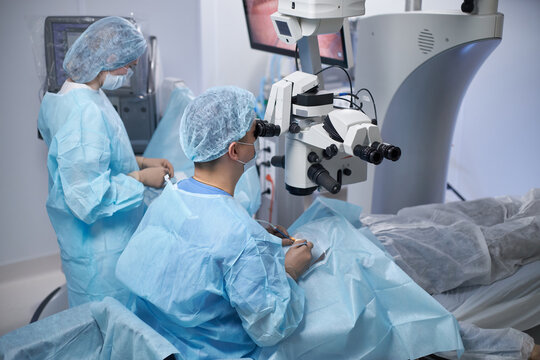 Middle aged man performing surgery using operating microscope while woman assisting in modern operating room with patient lying on table