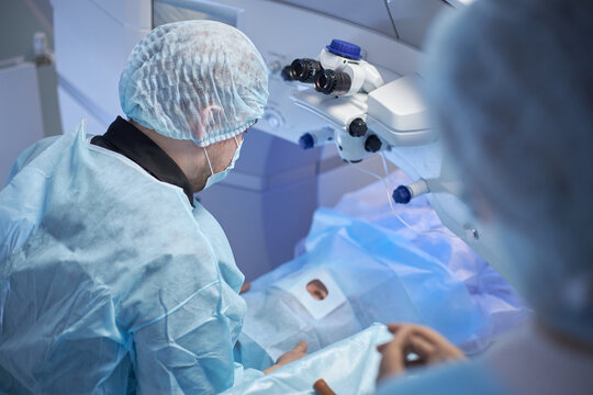 Middle aged male surgeon performing eye surgery on patient in operating room, using advanced ophthalmic microscope, medical staff assisting during delicate procedure