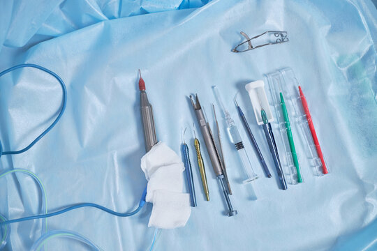 Surgical instruments and medical devices arranged on sterile blue drape in operating room setting, including scalpel, syringe, forceps, needle holder, and electrosurgical pencil