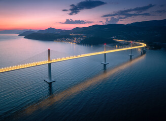 Aerial view of modern Peljesac bridge and blue sea at night. Croatia. Bridge with illumination, mountains, town, reflection and purple sky at sunset. Top drone view of road, street lights at twilight
