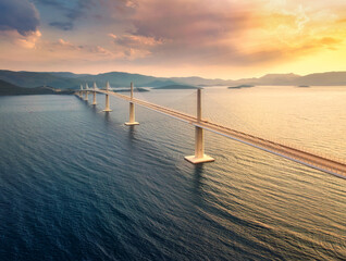 Aerial view of modern Peljesac Bridge and blue sea at colorful sunset in summer. Croatia. Top drone view of bridge during golden hour, mountains, reflection in water and orange sky. Architecture