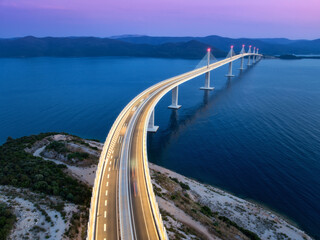 Aerial view of modern Peljesac bridge and blue sea at night. Croatia. Bridge with illumination, mountains, reflection and purple sky at sunset. Top drone panoramic view of road, street lights at dusk