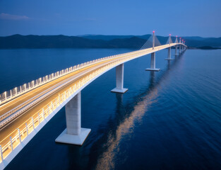 Aerial view of modern Peljesac bridge and blue sea at night. Croatia. Bridge with illumination, mountains, reflection and purple sky at sunset. Top drone panoramic view of road, street lights at dusk