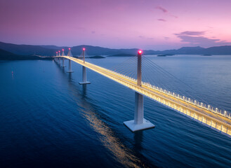 Aerial view of modern Peljesac bridge and blue sea at night. Croatia. Bridge with illumination, mountains, reflection and pink sky at sunset. Top drone panoramic view of road, street lights at dusk