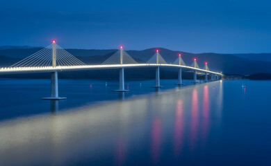 Aerial view of modern Peljesac bridge and blue sea at night. Croatia. Bridge with illumination, mountains, blurred reflection in water. Panoramic view of road, street lights at dusk. Architecture