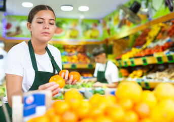 Smiling female seller standing at counter and lay out ripe tangerines in grocery supermarket