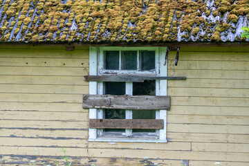 Old Wooden House Window Barricaded with Planks – Moss-Covered Roof and Rustic Yellow Wall Texture