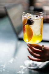 Close-up of a hand holding a glass of refreshing iced drink with fruit and berries, against a blurred background. Vibrant summer beverage with citrus and sparkling bubbles