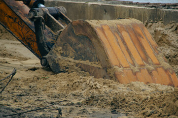 A close-up view of an excavator's bucket digging into sandy soil at a construction site.