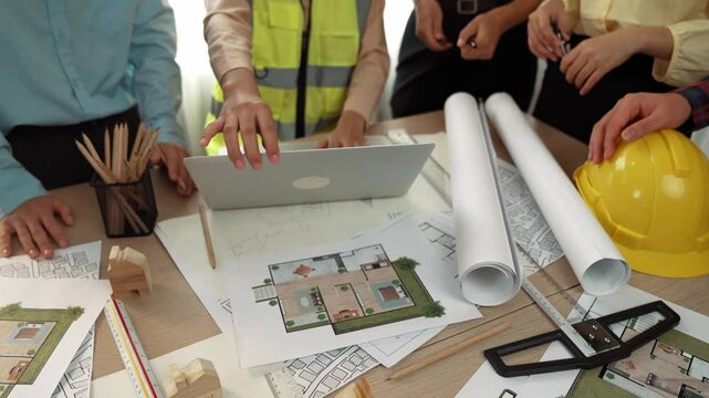 Architect and safety engineer discussion about building blueprint displaying on laptop on table with architectural document and wooden block scatter around at modern office. Closeup. Delineation.
