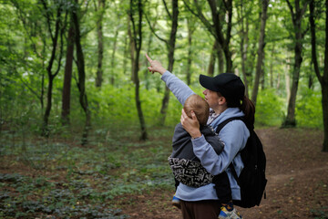 Mother carrying baby in baby carrier during forest trip and pointing at bird sitting on tree. Woman with backpack walking in green forest enjoying time outdoors with family.