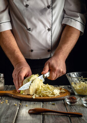 A chef skillfully slices fresh cabbage on a wooden cutting board while surrounded by various kitchen utensils and ingredients in a dimly lit environment