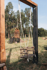  Rustic baby swing made of wood and rope in a backyard playground, with a handmade playhouse and trees in the background on a sunny day
