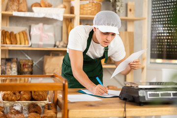 Near counter in bakery, guy seller reads report document, writes data in declaration, counts...