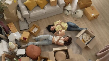 Smiling lesbian couple lying on the floor while spending time together. Top down view of girl lover reading a book while lying at living room surrounded with cardboard box in moving day. Myrmidon.