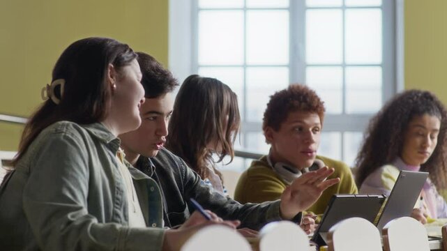 Stab shot of five multiethnic gen Z peers chatting sitting at desk with digital gadgets in uni auditorium