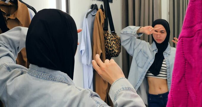 Confident young muslim woman in hijab standing near mirror, selecting stylish denim jacket while exploring clothing options inside retail boutique fitting room. concept of freedom for Muslim women