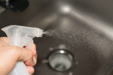 A hand presses a cleaning spray bottle, releasing a fine mist of solution into a stainless steel kitchen sink, capturing the motion of droplets in mid-air, representing cleanliness and disinfection