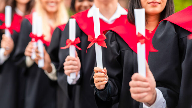 Unrecognizable international group of male and female graduators wearing graduation robes showing their diplomas, closeup. High school, university college graduation concept