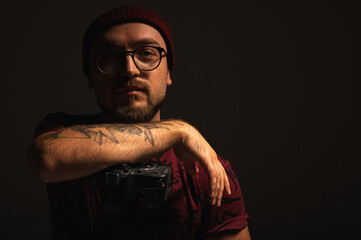 Portrait of a stylish young male photographer with tattoos and glasses, wearing a red beanie,...