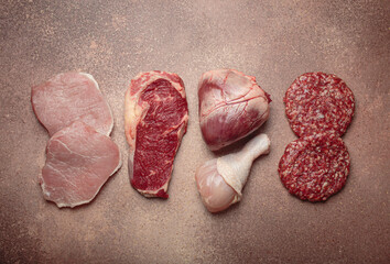 Selection of assorted raw meat cuts for zero carb carnivore diet: uncooked beef steak, pork, ground meat patty, heart, and chicken legs on beige stone background top view