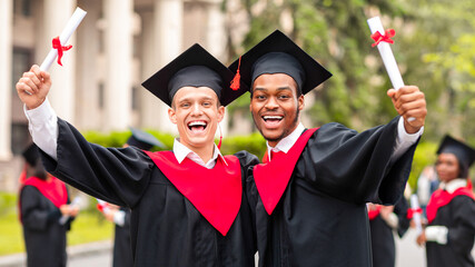 Happy guys students posing together next to university, wearing graduation costumes and showing...