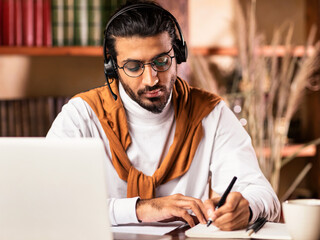 Indian Student Guy Taking Notes Learning At Laptop Doing University Homework Online Studying...