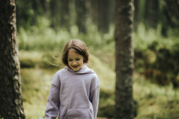 A young girl in a lavender hoodie and pink pants walks through a mossy forest, holding a plastic jar, possibly collecting insects or exploring nature.