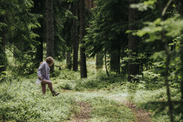A young girl in a lavender hoodie and pink pants walks through a mossy forest, holding a plastic jar, possibly collecting insects or exploring nature.