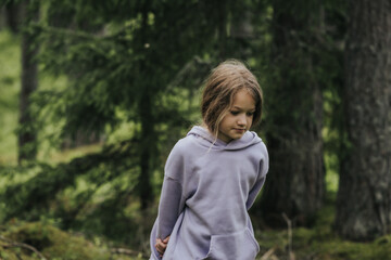 A young girl in a lavender hoodie and pink pants walks through a mossy forest, holding a plastic jar, possibly collecting insects or exploring nature.