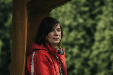 A woman in a red outdoor jacket leans against a large wooden structure with a calm expression, surrounded by a lush green forest background.