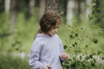 A young girl in a lavender hoodie and pink pants walks through a mossy forest, holding a plastic jar, possibly collecting insects or exploring nature.