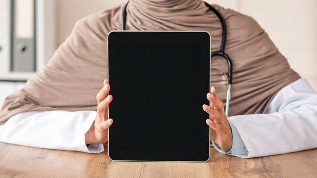 Cropped of female doctor muslim woman in hijab showing digital tablet with blank screen, mockup, copy space. Unrecognizable doctor using modern technologies in her job, working and training online