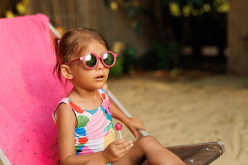 Funny cute girl on summer vacation. The child has fun near the pool. Cute baby girl in a colorful swimsuit and sunglasses is resting.