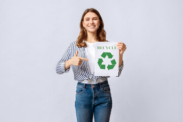 Happy Young Eco-Volunteer Lady Holding Placard With Green Recycle System Icon With Circulating Arrows And Pointing On It With Finger, Smiling Environmental Activist Woman Standing On Light Background