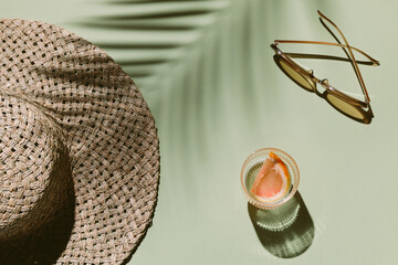 Summer flat lay with straw hat, sunglasses and glass of water with grapefruit slices on green background with palm leaf shadow, sun and sunlight. Vacation, holiday, summer creative minimal concept.