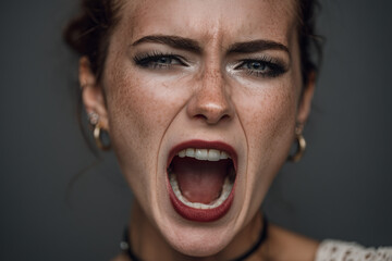 Emotional young woman with freckles expressing intense feelings while shouting against a dark background in a close-up portrait, showcasing her vibrant personality and bold makeup