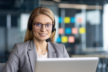 A friendly blonde woman with glasses wears a headset, smiling at the camera while working on a laptop in an office setting.