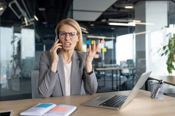 An angry businesswoman in a modern office is on a phone call, looking frustrated and stressed. She is gesturing with her hand and wearing glasses.