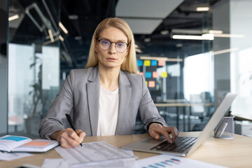 A professional businesswoman in a modern office setting, working on a laptop and taking notes, with...
