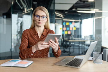 A businesswoman with glasses works with a tablet and laptop in a modern office setting.