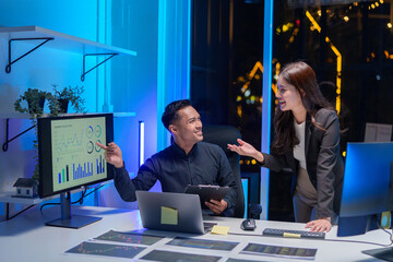 Two Asian business partners discussing work while using laptops in an office at night.