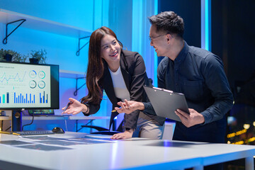 
Young Asian business team working late into the night in a well-lit office, collaborating on a project using laptops.